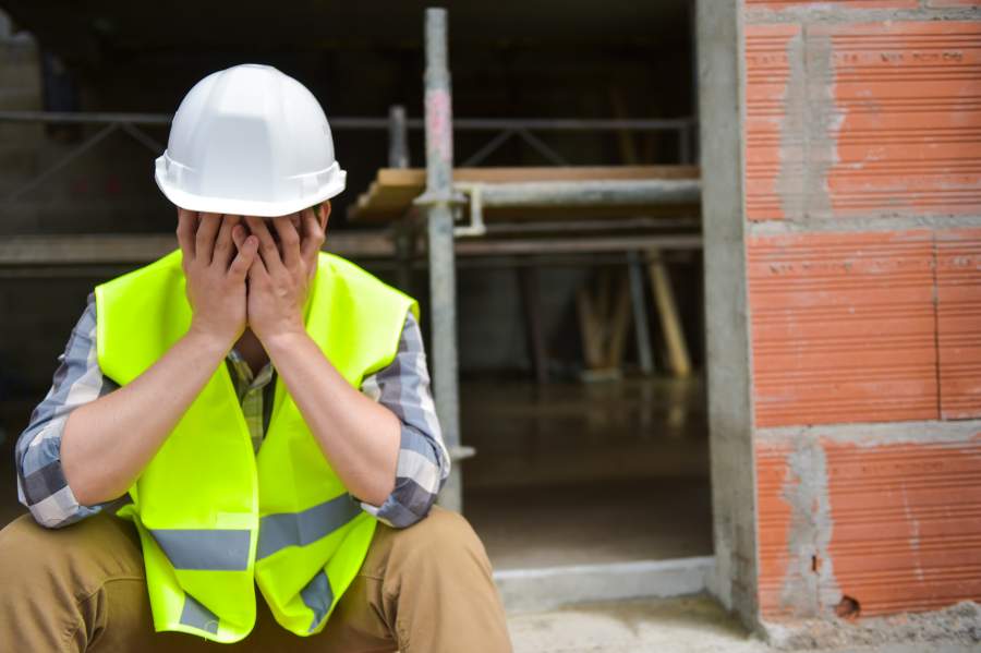 Un travailleur fatigué, frustré, portant un casque de sécurité blanc et un gilet de sécurité lui frappe les mains devant le visage