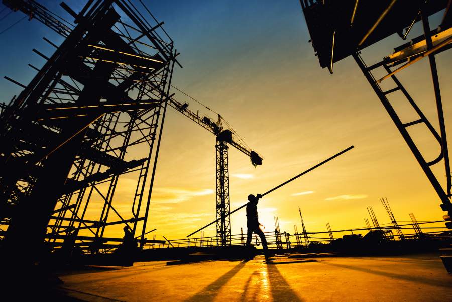 La silhouette d'un employé portant une longue barre de fer au-dessus de son épaule sur le chantier de construction, avec le coucher du soleil en arrière-plan.