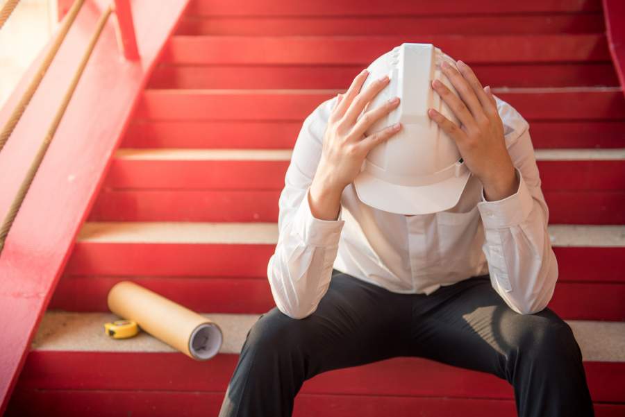 Un employé stressé et fatigué avec un casque de sécurité blanc s'assoit sur un escalier rouge et touche sa tête